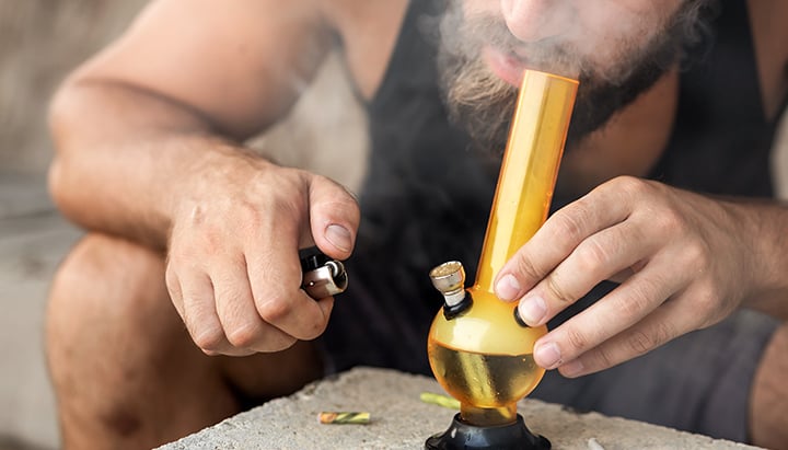 Persona fumando hachís de un bong de vidrio, exhalando humo espeso.